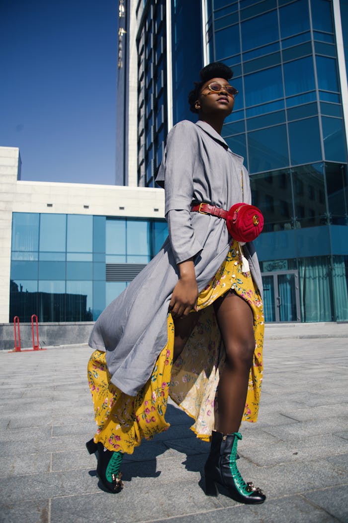 A fashionable woman in a vibrant yellow dress and grey coat posing confidently in an urban environment.