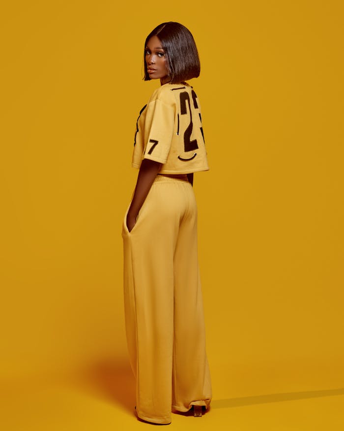 Fashionable woman wearing yellow clothing stands against a vibrant yellow background, turning slightly.